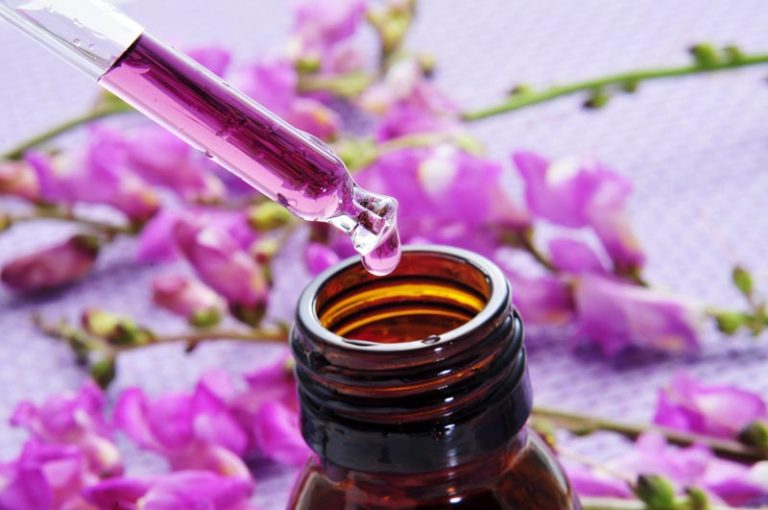 Eine Pipette füllt Trpfen in ein Flasche, im Hintergrund violette Blüten.